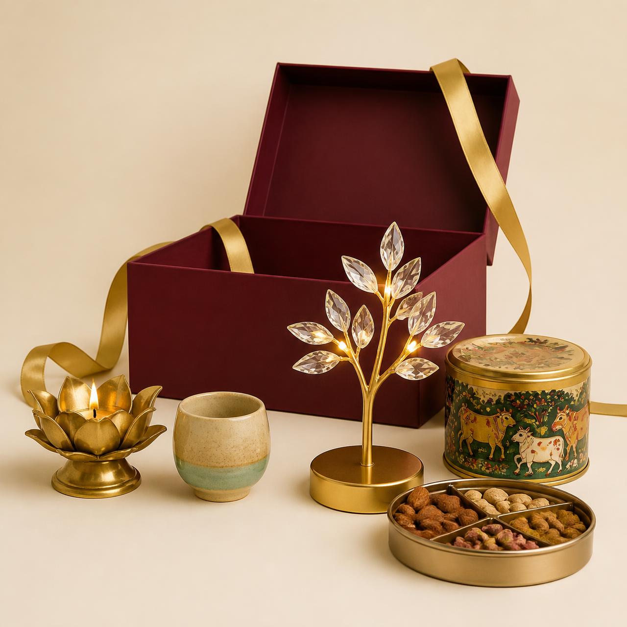 An open burgundy gift box surrounded by a brass lotus diya, ceramic cup, crystal leaf lamp, Pichwai-print container and a dry fruit box